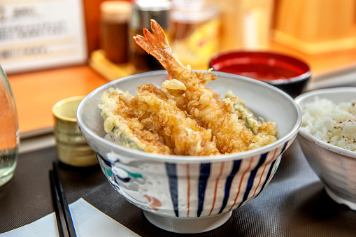 Japanese deep fried prawn and vegetables tempura, in a bowl with rice. Chopsticks, saki, ondiments and sauces in the background