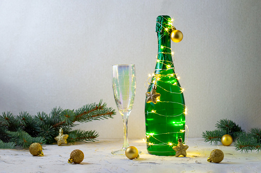 a bottle of green champagne wrapped in a glowing garland like a Christmas tree and glass. New year concept with gold decor on white background