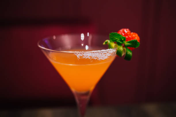 Closeup of glass of alcohol cocktail decorated with mint and strawberry  placed on wooden table against blurred red wall in bar