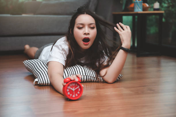 Young asian woman open mouth yawning and reaching over to turning off alarm clock while lying on the floor in living room at home. Young asian woman open mouth yawning and reaching over to turning off alarm clock while lying on the floor in living room at home. woman lying on the floor isolated stock pictures, royalty-free photos & images