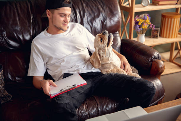 istock Young man sitting on sofa at home with his cute dog. Cozy office workplace, remote work, online, e-learning concept. 1288836349