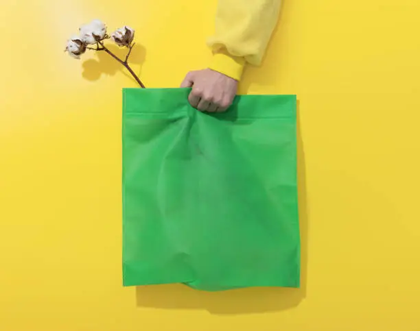 Hand holds green cotton bag and Cotton flowers on yellow background