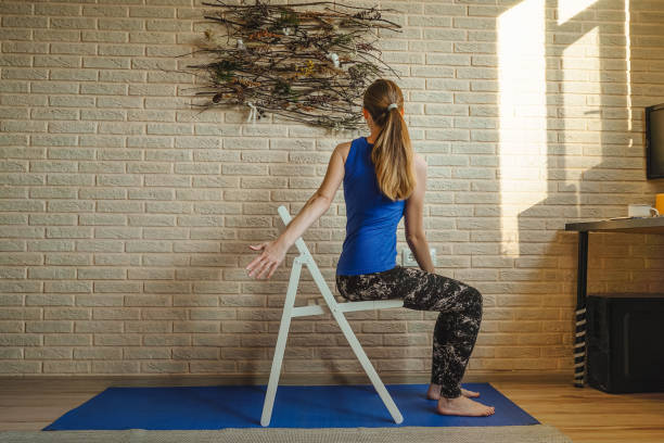Woman doing yoga at home. Woman doing yoga at home. Blue mat and undershirt. Twisting pose with a chair, sunny day. Natural panels on the brick wall, sustainable design. Attending to mental health. twist pose stock pictures, royalty-free photos & images