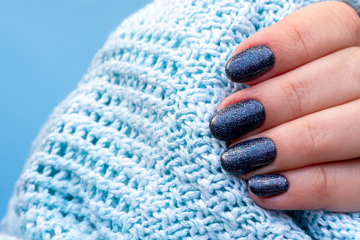 Female hand with knitted sweater fabric with beautiful manicure - dark blue glittered nails on blue background. Selective focus. Closeup view