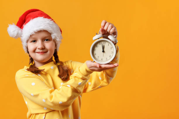 vigilia di natale. ritratto di una bambina carina con un cappello di babbo natale. il bambino tiene in mano una sveglia su sfondo giallo. - clock hand eve time white foto e immagini stock