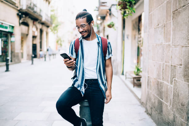 istock Smiling african american trendy dressed hipster guy in eyewear recreating outdoors using smartphone and sound application for enjoy music, positive dark skinned male listening audiobook on street 1288875990