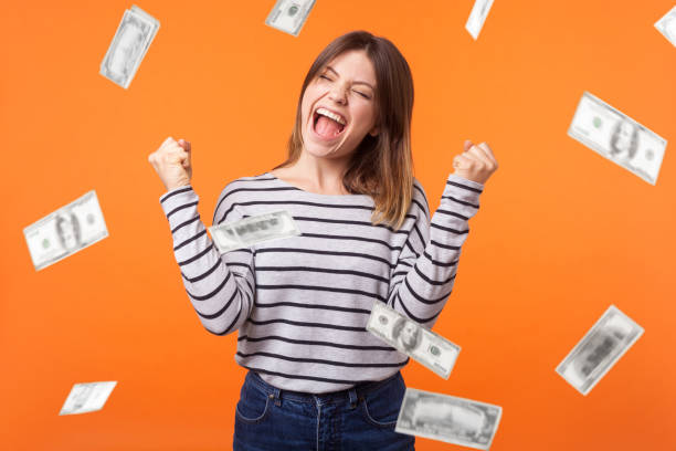 Money rain. Yes I did it! Portrait of joyous winner, young woman in casual shirt standing with clenched fists and closed eyes, celebrating victory and richness. Money rain. Yes I did it! Portrait of joyous winner, young woman in casual shirt standing with clenched fists and closed eyes, celebrating victory and richness. indoor isolated on orange background super money stock pictures, royalty-free photos & images