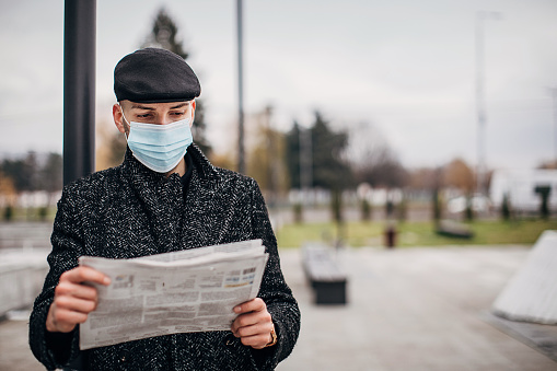 One man, handsome modern young man with protective face mask standing on the street alone in city, he is reading newspaper.