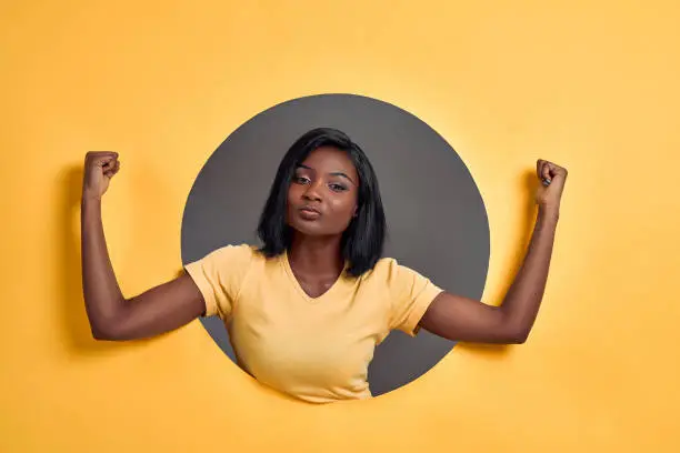 Photo of Woman raises hands and shows muscles, demonstrates her strength, wears yellow t-shirt, says: I am winner or champion. People, triumph concept. Feminism.
