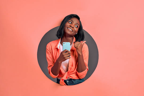 jonge afro amerikaanse vrouw die gebruikend smartphone over geïsoleerde achtergrond sms'te die en met duim omhoog aan de kant met gelukkig gezicht toont dat glimlacht toont. ruimte kopiëren. - phone hand thumb stockfoto's en -beelden