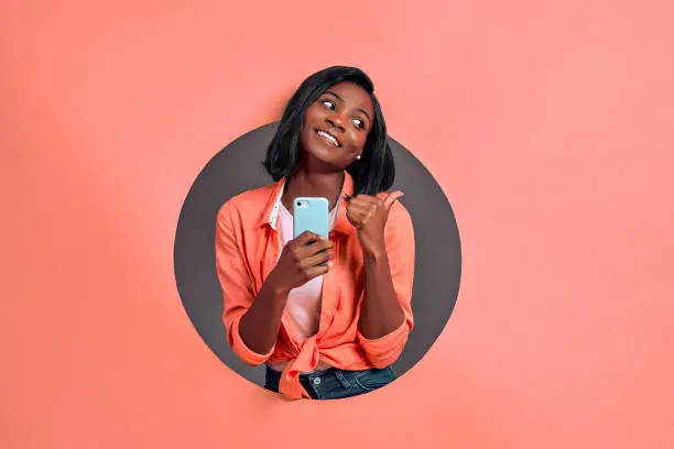 Photo of Young afro american woman texting using smartphone over isolated background pointing and showing with thumb up to the side with happy face smiling. Copy space.