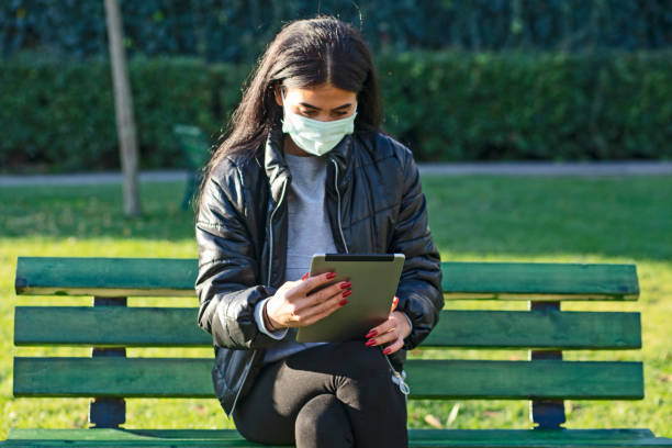 istock Young woman with face mask sitting on a bench in a public park using digital tablet 1288885290