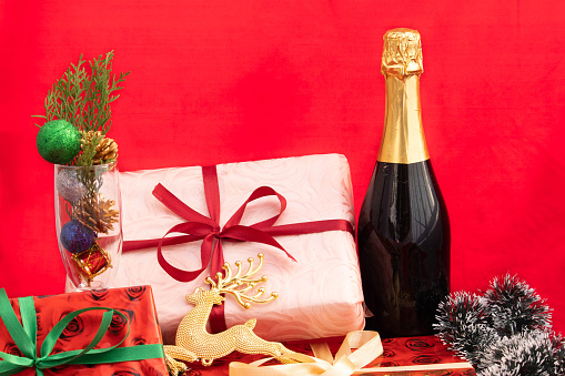 Lots Of Colorful Packed Christmas Gift Boxes Tied With Shining Ribbons And Decoration Items In Wine Glass And Closed Pack Champagne Bottle Neck Wrapped With Golden Paper At Top On Red Background