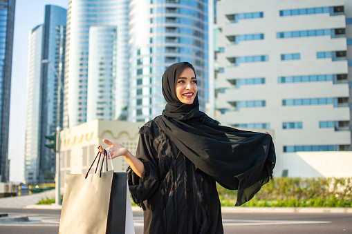 Hermosa joven mujer musulmana árabe con ropa de abaya sosteniendo bolsas de compras y caminando por la calle de la ciudad. Tiempo de compras. Modernos rascacielos en el fondo. photo