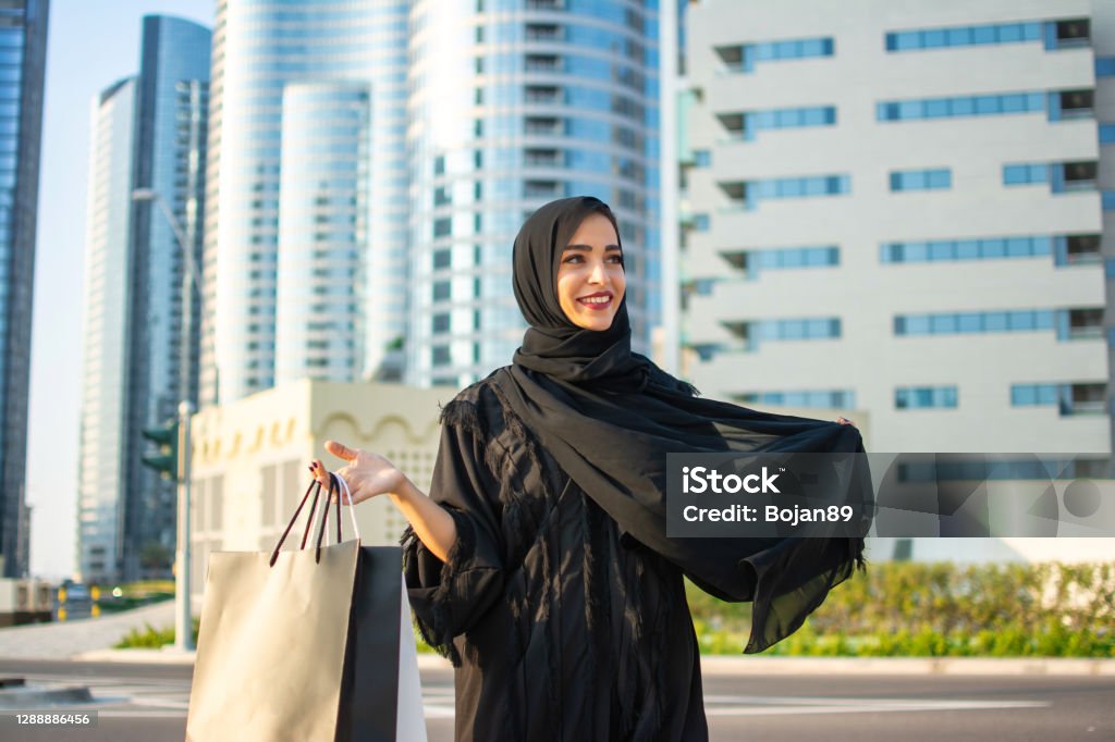 Beautiful young arab muslim woman in abaya clothes holding shopping bags and walking on the city street. Shopping time. Modern skyscrapers in the background. Women Stock Photo