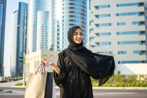 Photo of Beautiful young arab muslim woman in abaya clothes holding shopping bags and walking on the city street. Shopping time. Modern skyscrapers in the background.