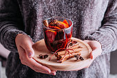Woman in grey winter sweater holding mulled wine with oranges and spices on tray, cozy winter lifestyle closeup