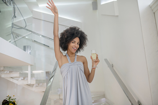 Celebration, emotion. Cheerful young woman in cocktail dress with glass on stairs in beautiful room