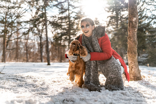 Beautiful young woman with sunglasses and scarf crouching in nature on snow and embracing her dog