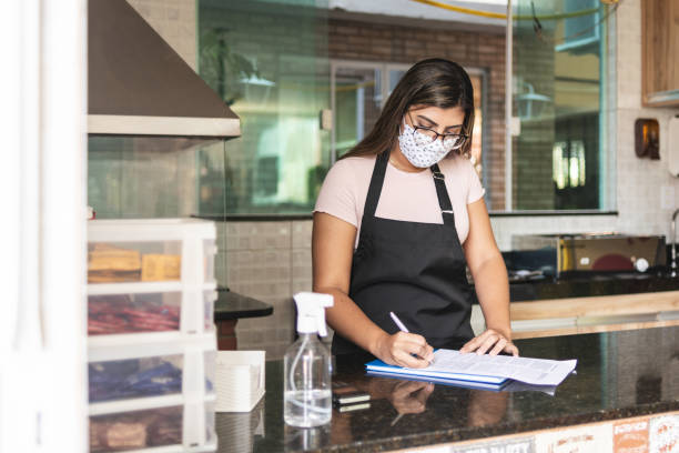 istock Young waitress or owner writing in a note pad at store counter, wearing face mask 1288896842