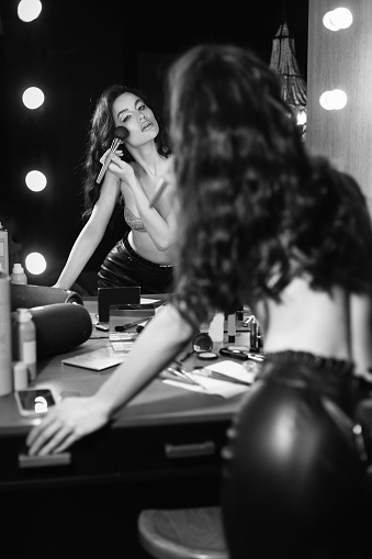 A beautiful girl in underwear and a leather skirt is making up in front of the mirror. A chic model with makeup and hair is going to a party.