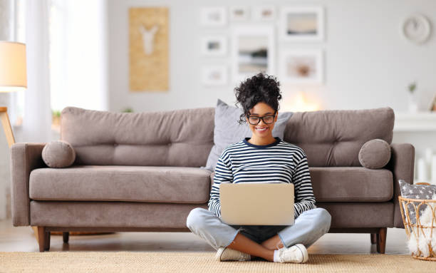 Joyful relaxed ethnic woman using laptop with interest at home Cheerful young ethnic female freelancer in glasses and casual clothes focusing on screen and interacting with laptop while sitting alone on floor in light modern living room netbook stock pictures, royalty-free photos & images