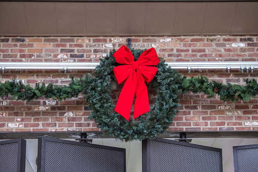 An outdoor festive Christmas green wreath with a bright red bow and garlend against a red brick wall