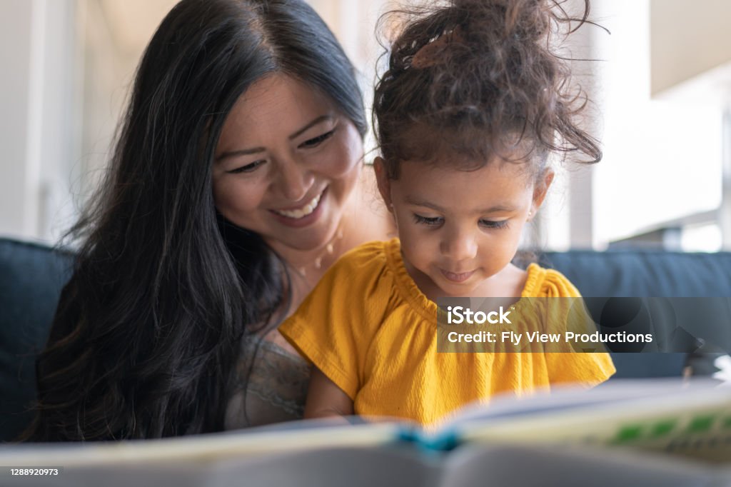 Adorable mixed race toddler reading book with her mother A loving mother of Asian descent sits on the couch at home and reads a storybook to her preschool age daughter. The child is sitting on her mother's lap and looking at the book intently. Reading Stock Photo
