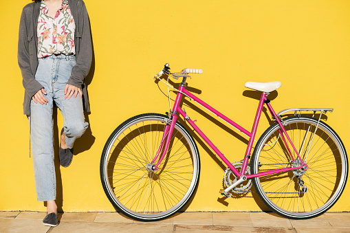 unrecognizable young woman leaning against a colorful yellow wall next to her pink retro bike, concept of active lifestyle and sustainable mobility, copy space for text
