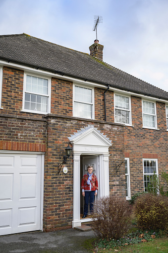 Caucasian woman in late 70s wearing casual clothing and standing inside open front door of home in West Sussex looking at camera with contented expression.