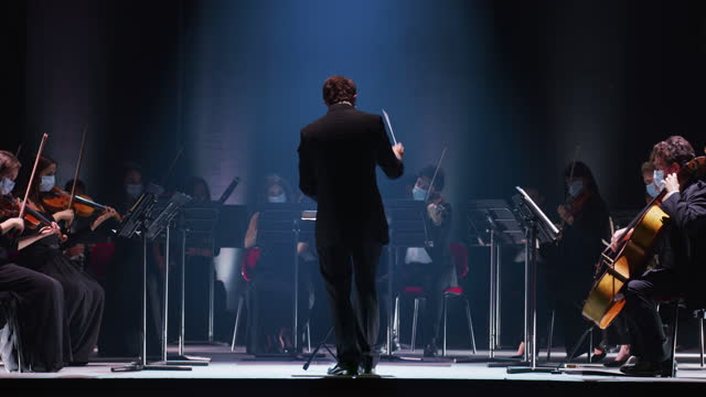 Cinematic shot of conductor directing symphony orchestra with performers with medical protective masks playing violins, cello and trumpet on classic theatre