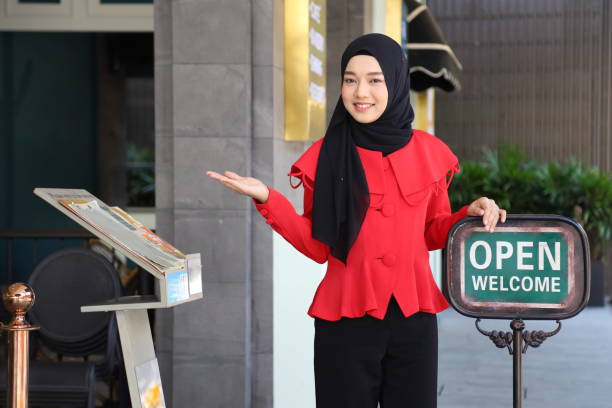 signora musulmana in piedi fuori dal ristorante halal con cartello aperto per accogliere il cliente - hotel greeting welcome sign service foto e immagini stock