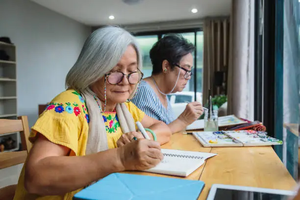 Photo of Asian senior woman friends enjoying drawing with pen while setting in living room at home, Hobby and relaxing concept