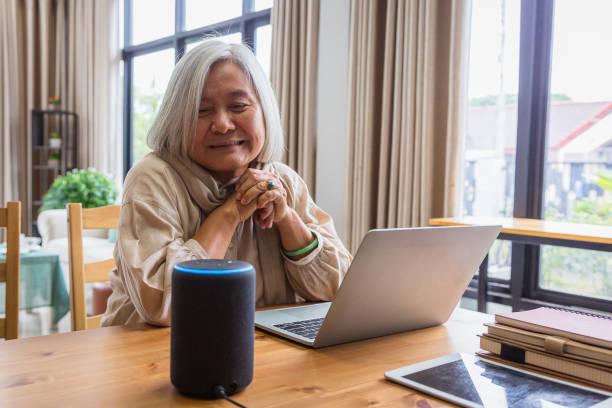 asiatische seniorinnen arbeiten mit laptop-computer und mit intelligenten lautsprechern, während sie im wohnzimmer zu hause - smarthome stock-fotos und bilder