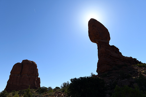 Balanced Rock, carved in sandstone by wind and rain erosion, stands still despite gravity. Pictured against the sun on a beautiful, sunny day. Arches National Park, Utah, USA. Jet airplane cuts the blue sky.