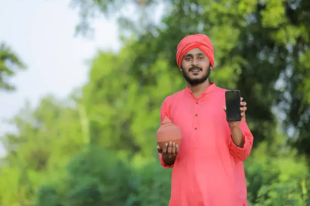 Photo of Young indian farmer holding clay piggy bank and showing smart phone in hand