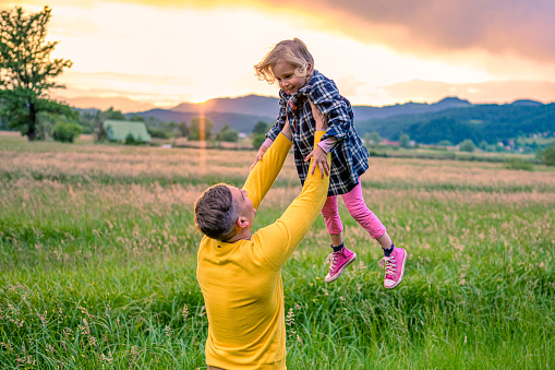Smiling father lifting his daughter into the air on a meadow at sunset.