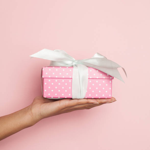 istock Minimal background composition with hand holding pink gift with white silky ribbon on pastel pink background 1288961852