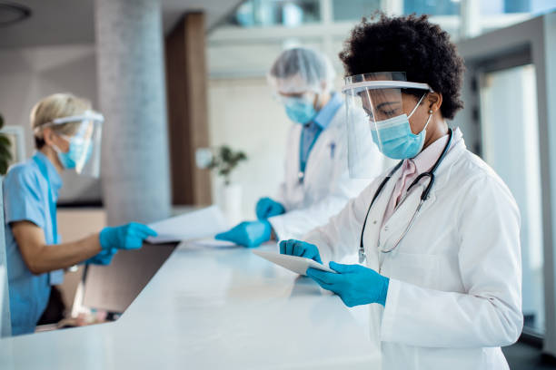 African American doctor working on touchpad at reception desk in the hospital during coronavirus pandemic. Black female doctor using digital tablet while standing at reception desk at medical clinic and wearing face shield and mask due to COVID-19 pandemic. protective workwear stock pictures, royalty-free photos & images