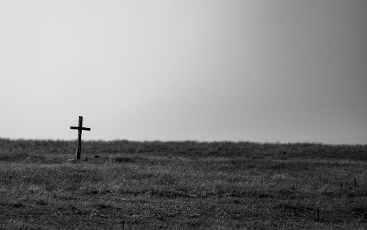 Black and white image of an old wooden Christian cross standing on a barren grass plain.