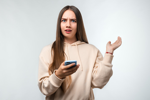 Portrait of an indignant girl with long chestnut hair, dressed in casual clothes, using mobile phone standing isolated over white background