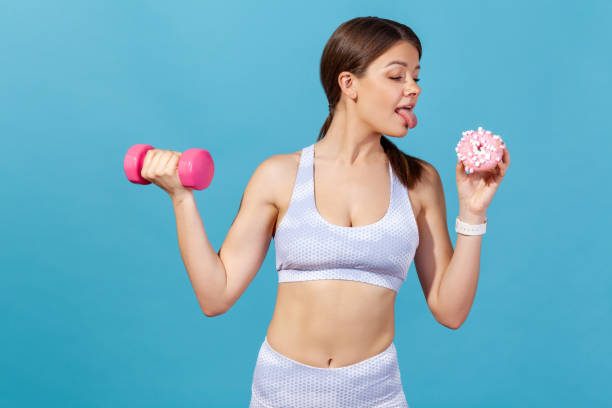 Athletic woman in white sports tights and top holding dumbbell and donut, showing tongue going to lick it, keeping diet going for sport Athletic woman in white sports tights and top holding dumbbell and donut, showing tongue going to lick it, keeping diet going for sport. Indoor studio shot isolated on blue background licking photos stock pictures, royalty-free photos & images