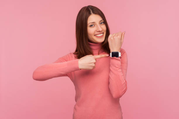 ピンクのセーターを着た茶色の髪の幸せなポジティブな女性は、歯の笑顔でカメラを見て手首に指を差し伸べています。時間を見て! - checking the time women impatient wristwatch ストックフォトと画像