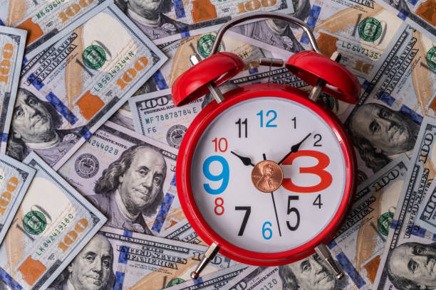 A red classic alarm clock lies against a background of 100 USD bills with a portrait of Benjamin Franklin A red classic alarm clock lies against a background of 100 USD bills with a portrait of Benjamin Franklin, symbolizing the saying "TIME is MONEY" wasting time stock pictures, royalty-free photos & images