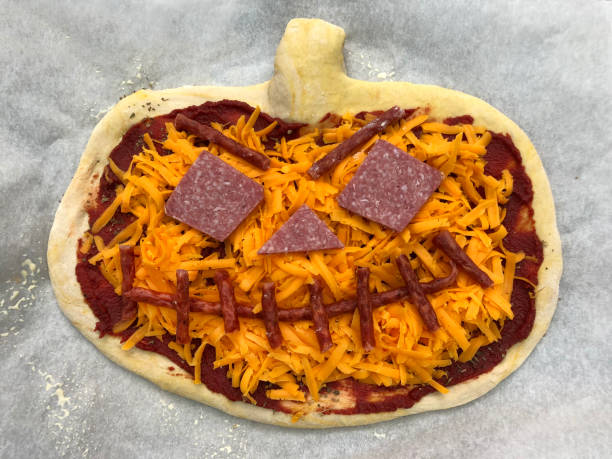 Stock photo showing elevated view of grease proof parchment paper lined baking tray containing a pizza cut like a Jack O'Lantern, topped with a rich tomato sauce, grated cheese and pepperoni and chorizo sausage. imagen de la idea de comida de fiesta temática de halloween jack o'lantern pizza cubierta con una rica salsa de tomate, queso rallado, pepperoni y salchicha de chorizo, en papel de pergamino a prueba de grasa forrado bandeja de hornear, vista elevada