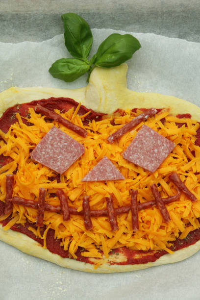 Stock photo showing elevated view of grease proof parchment paper lined baking tray containing a pizza cut like a Jack O'Lantern, topped with a rich tomato sauce, melted cheese and pepperoni and chorizo sausage. imagen de la idea de comida de fiesta temática de halloween jack o'lantern pizza cubierta con una rica salsa de tomate, queso derretido, pepperoni y salchicha de chorizo, en papel de pergamino a prueba de grasa forrado bandeja de hornear, vista elevada