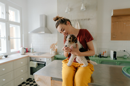 Photo of a young woman and her dog in the kitchen; the morning routine of a young woman and her pet. They are staying in quarantine during Covid 19 pandemic to avoid getting sick.