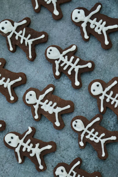 istock Full frame image of rows of homemade, people-shaped, chocolate biscuits on greaseproof parchment baking paper, cookies iced as skeletons with white icing, elevated view, Halloween themed party food 1288986143