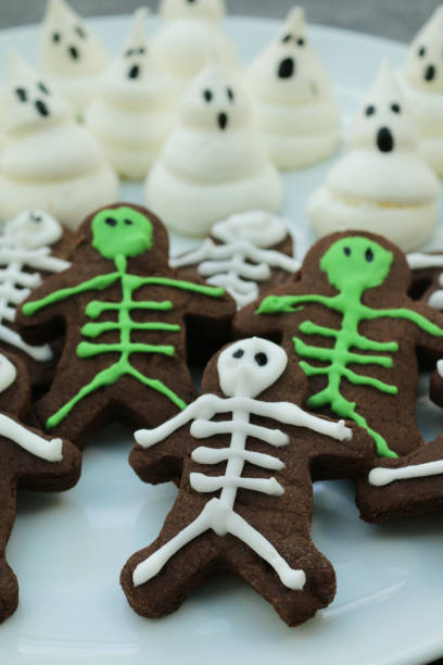 Stock photo showing elevated view of white plate containing freshly prepared, homemade people-shaped chocolate cookies decorated as skeletons with green and white icing besides cooked meringue peak ghosts. Halloween themed party food idea. imagen de primer plano de plato blanco de galletas caseras, en forma de personas, chocolate, galletas heladas como esqueletos con hielo verde y blanco al lado de los fantasmas pico merengue, se centran en el primer plano, comida de fiesta temática de hall