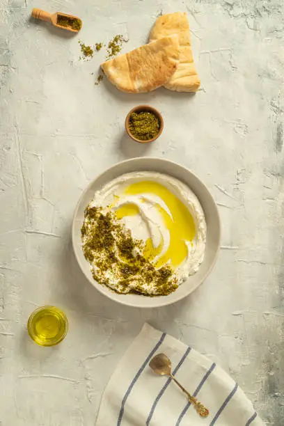Photo of Popular middle eastern appetizer labneh or labaneh, soft white goat milk cheese with olive oil, hyssop or zaatar, served with pita bread on a grey table, top view,
